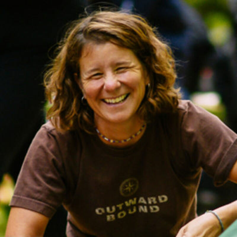 The image shows a woman with short, wavy brown hair smiling broadly. She is wearing a brown t-shirt with the words "OUTWARD BOUND" printed on it. She appears to be outdoors, possibly in a natural setting, and seems happy and relaxed. The background is blurred, suggesting a focus on the woman.